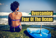 A woman sitting on the beach next to a surfboard with the words "Overcoming fear of the ocean"