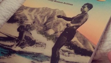 Book titled "Surfing: Historic images from Bishop Museum Archives" with a vintage photo of a person surfing in the ocean, with mountains in the background.